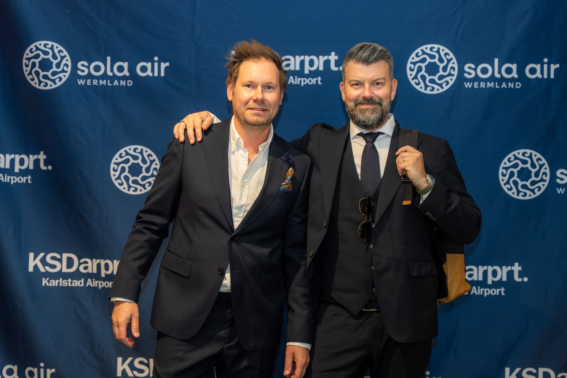 Två män i kostymer poserar vid en bakgrund av ett evenemang med varumärkeslogotyperna ”Sola Air” och ”KSDarpt”