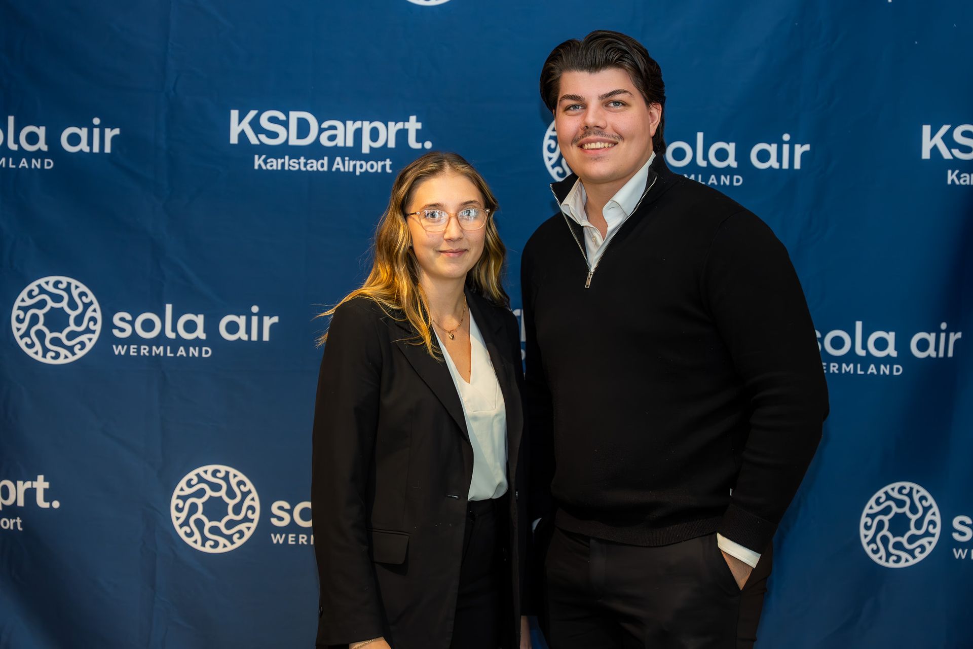 Två personer poserar framför en blå steg-och-repeat-bakgrund med Sola Air- och KSDart-logotyper
