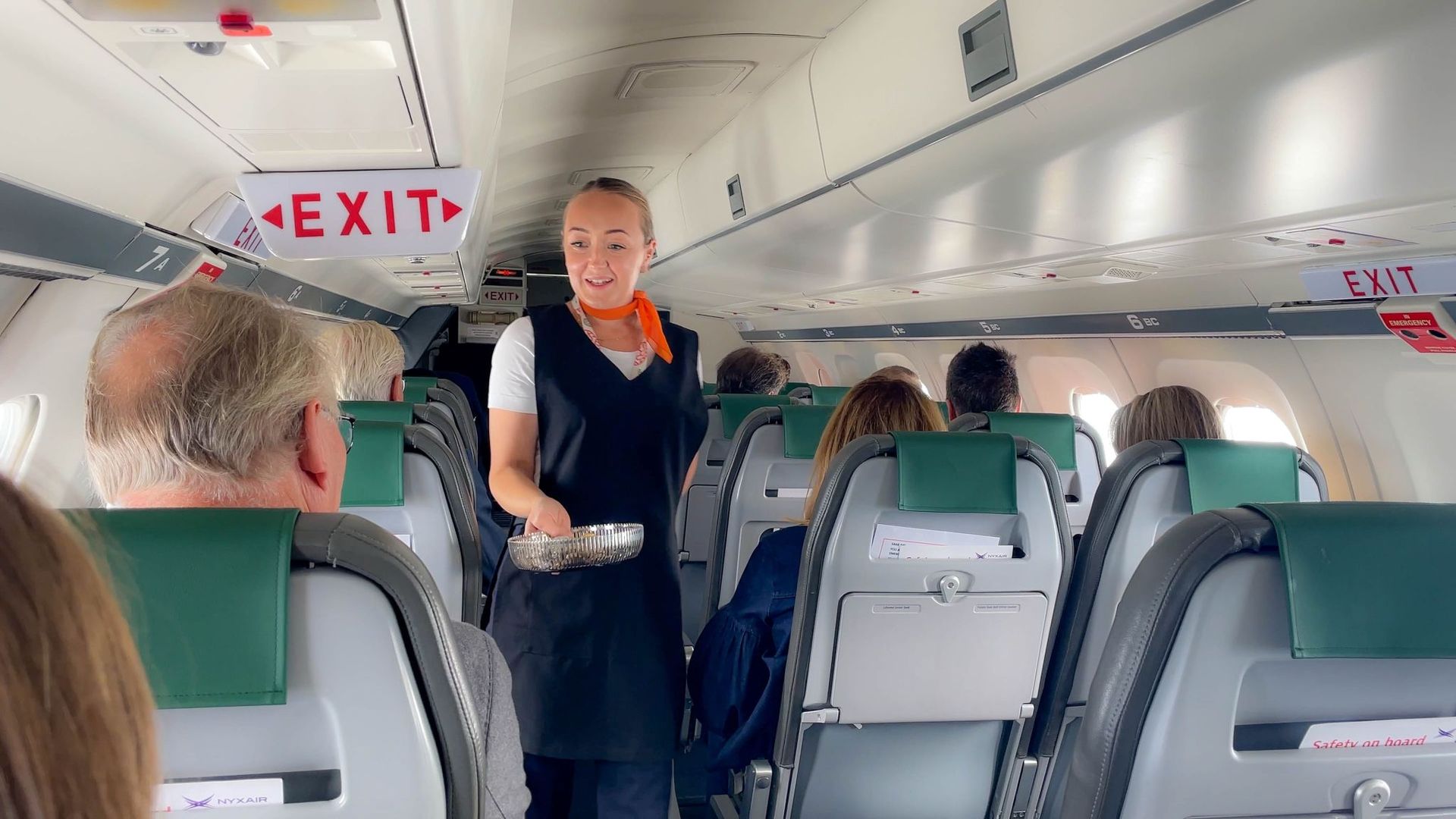 Flight attendant standing in an airplane aisle, serving passengers seated in green seats.