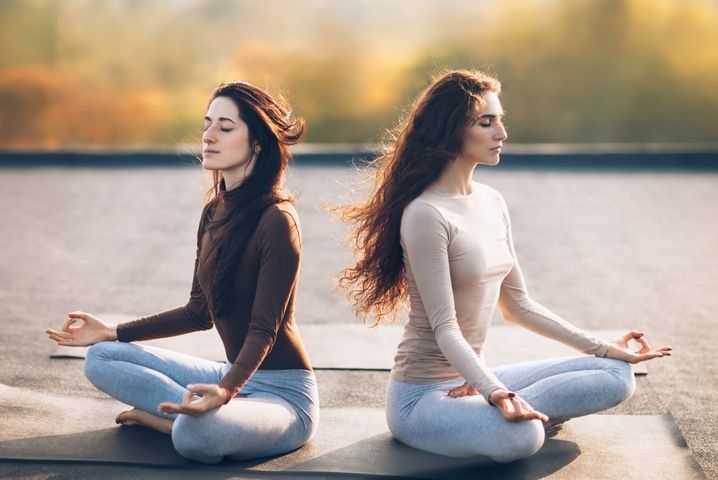 Two women meditating on a rooftop, eyes closed, hands in a mudra pose, wearing activewear.
