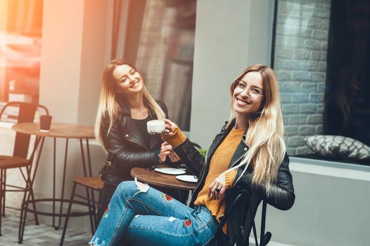 Two women with long blonde hair laughing, sitting at a cafe table, one holding a cup.