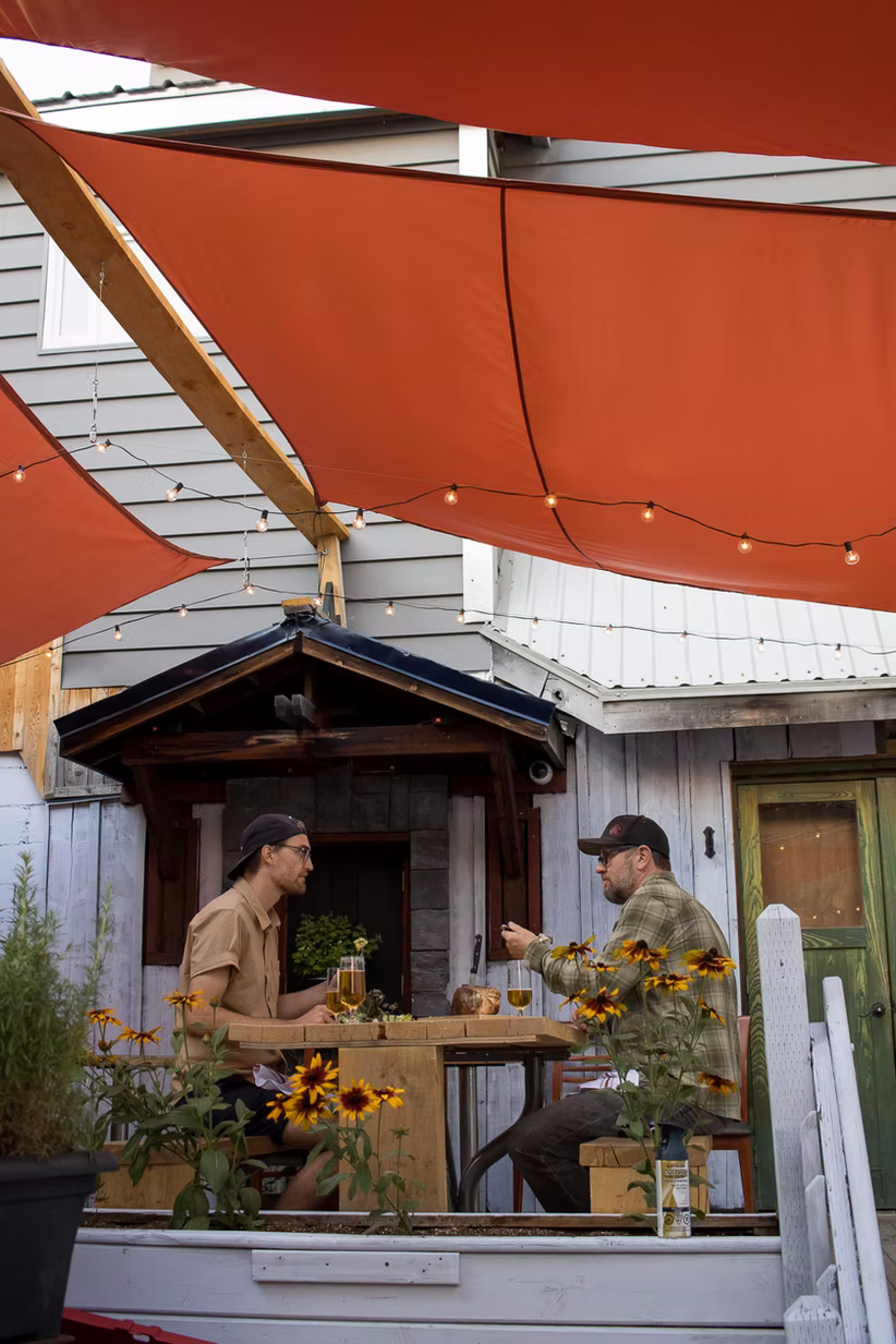 Two people seated at a table outdoors, under orange shade. Building in background, flowers in front.