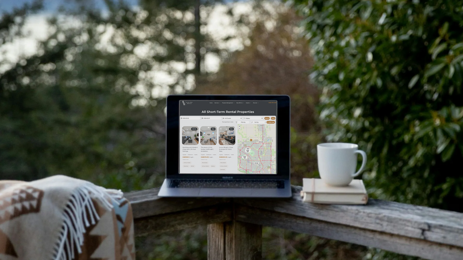 A laptop displaying a map and property listings sits on a wooden deck next to a coffee mug and a book, near leafy trees.