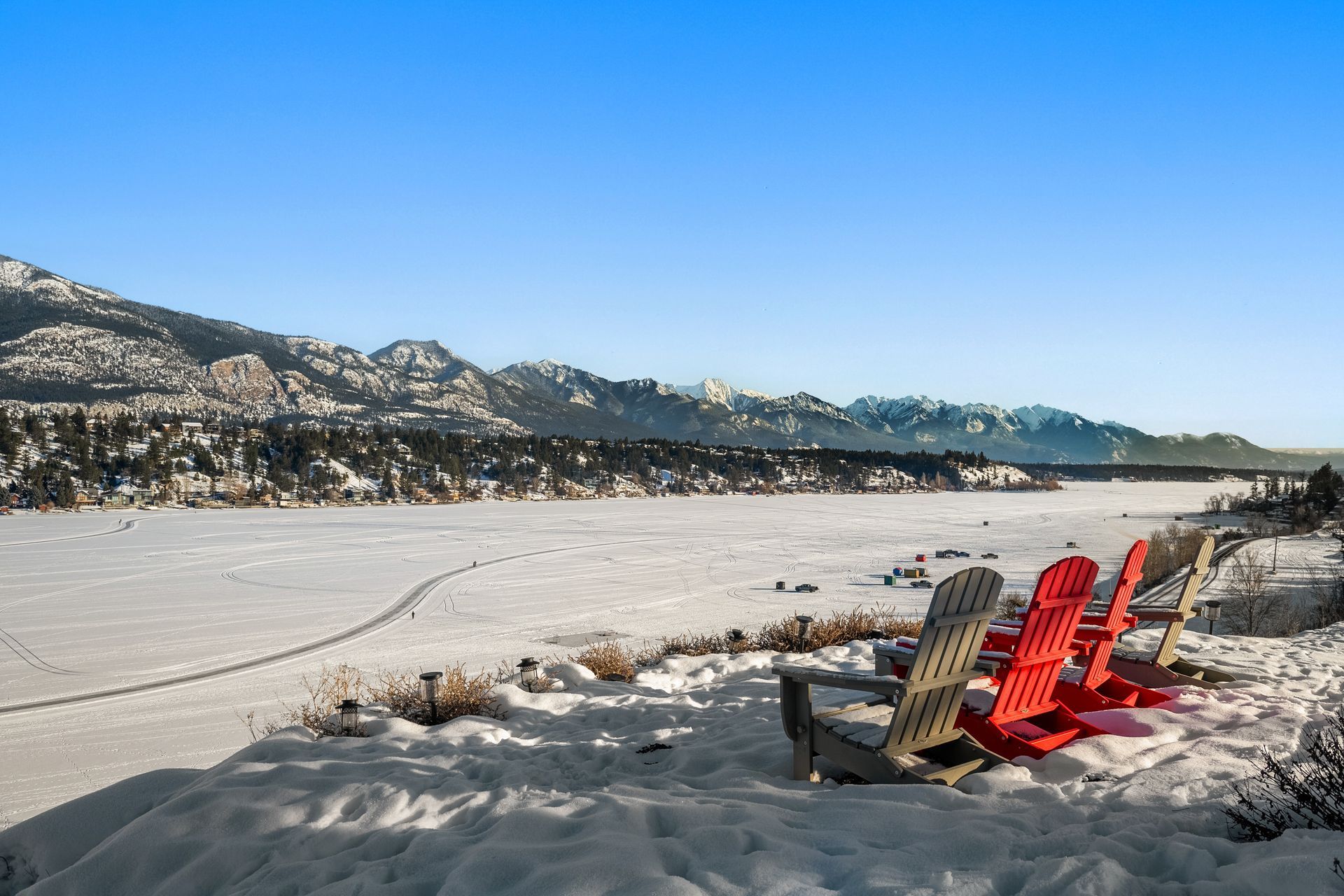 A row of red chairs are sitting in the snow near a lake.
