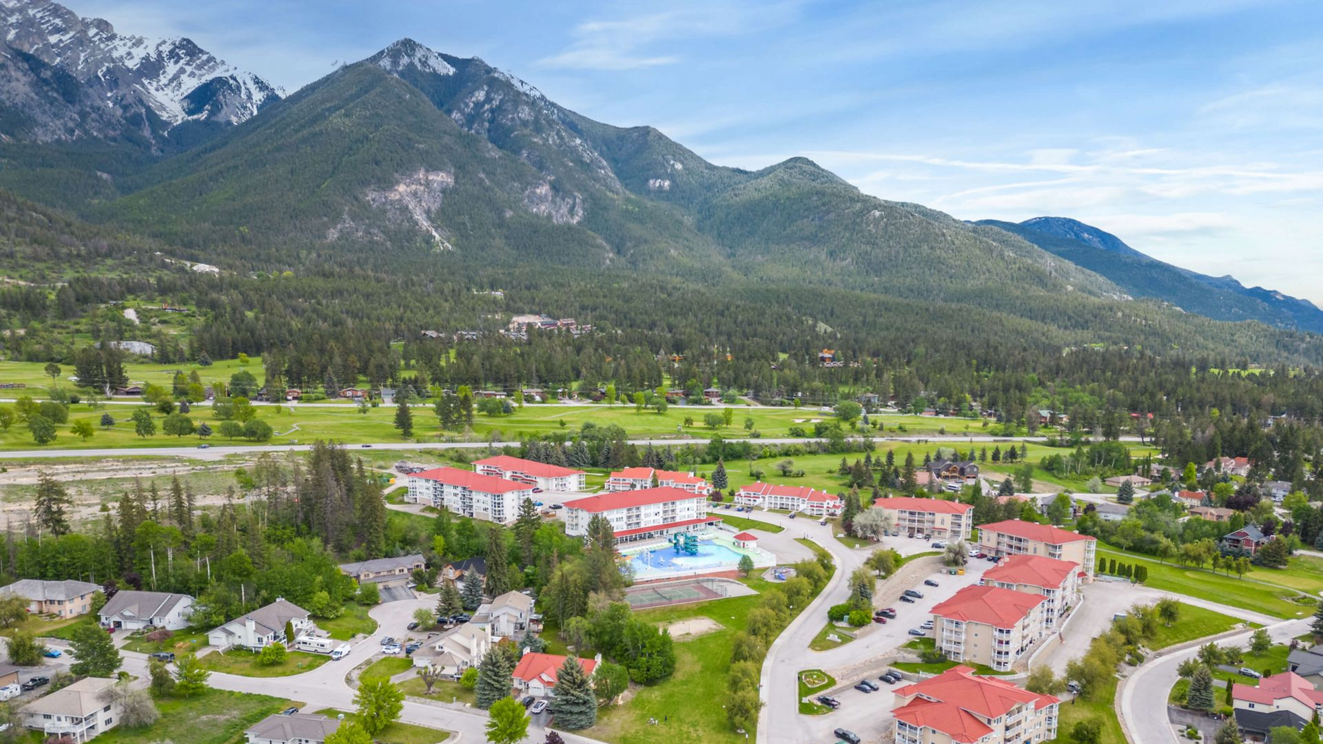 Aerial view of a mountain town with red-roofed buildings, green fields, and snow-capped peaks under a blue sky