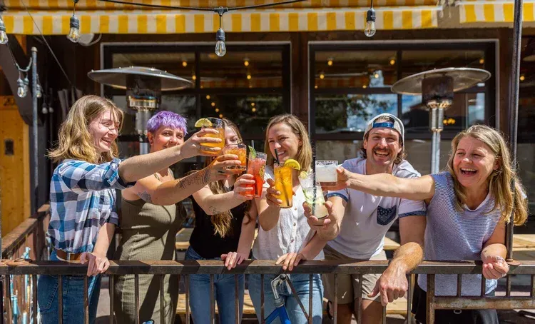 Group of people toasting cocktails on a patio. Yellow awning, outdoor heaters, and sunny day.