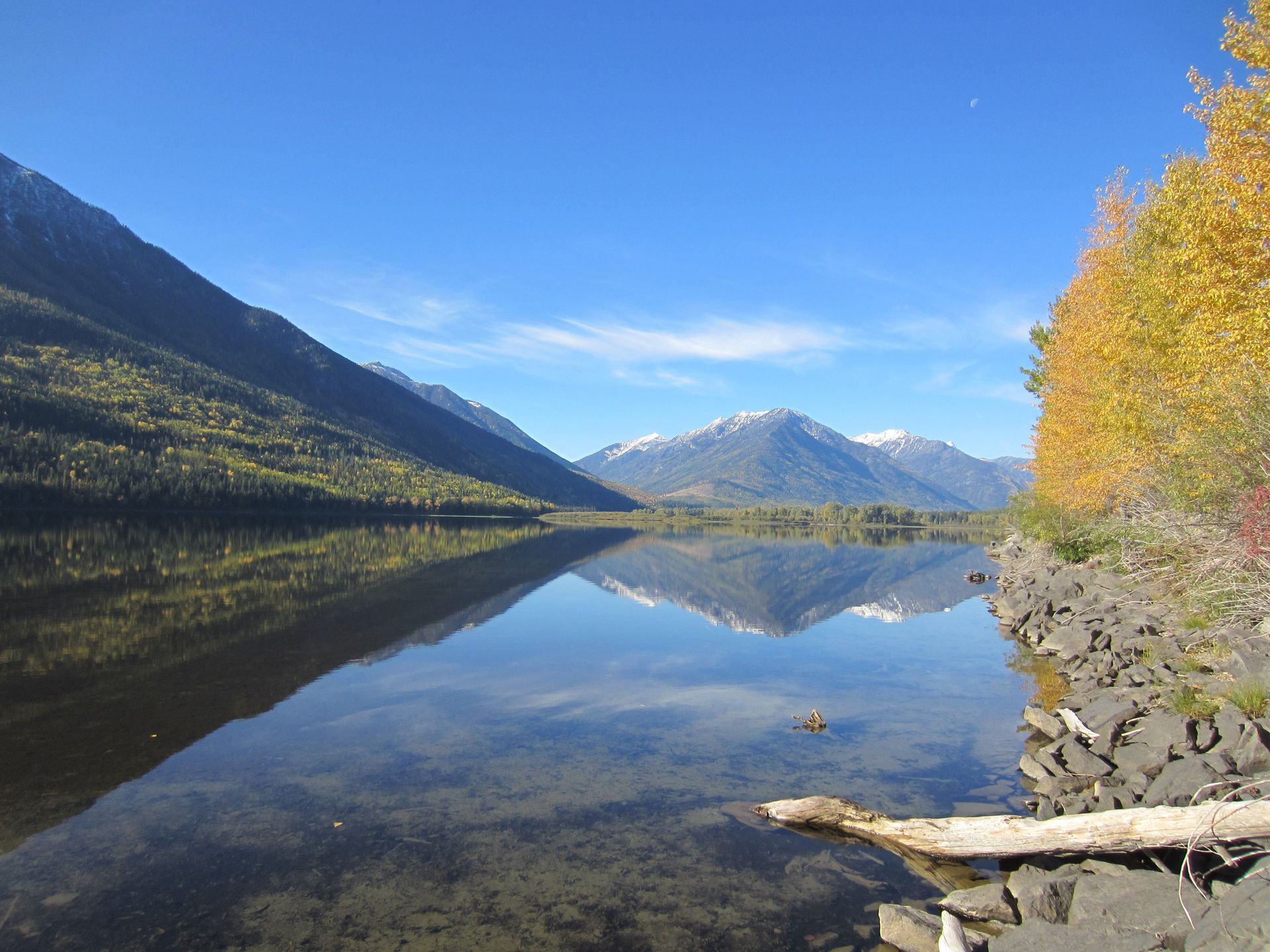 Calm lake reflects mountains and blue sky, with autumn trees on the right and rocky shore.