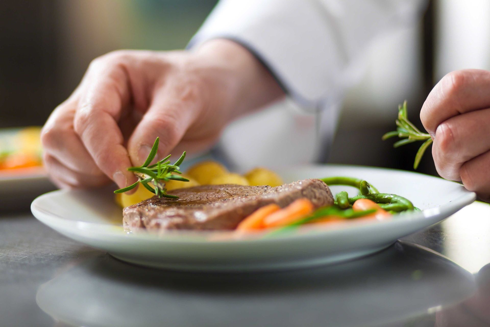 Chef garnishes plate with rosemary sprigs; steak, potatoes, and vegetables are plated.