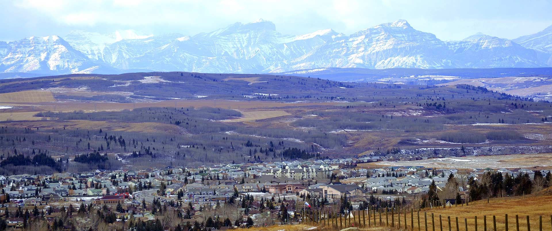 Snow-capped mountains and a town are seen from a field on a cloudy day.