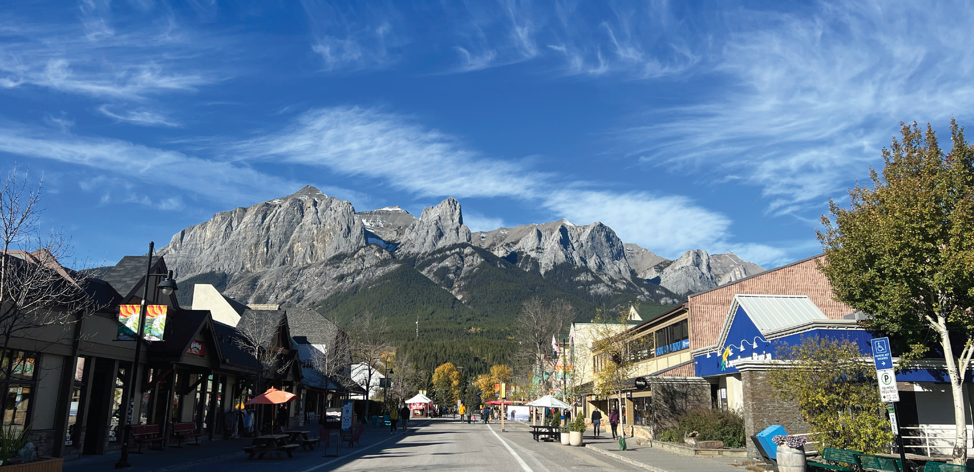 Mountain town street with colorful shops and trees, backed by rocky peaks under a blue sky
