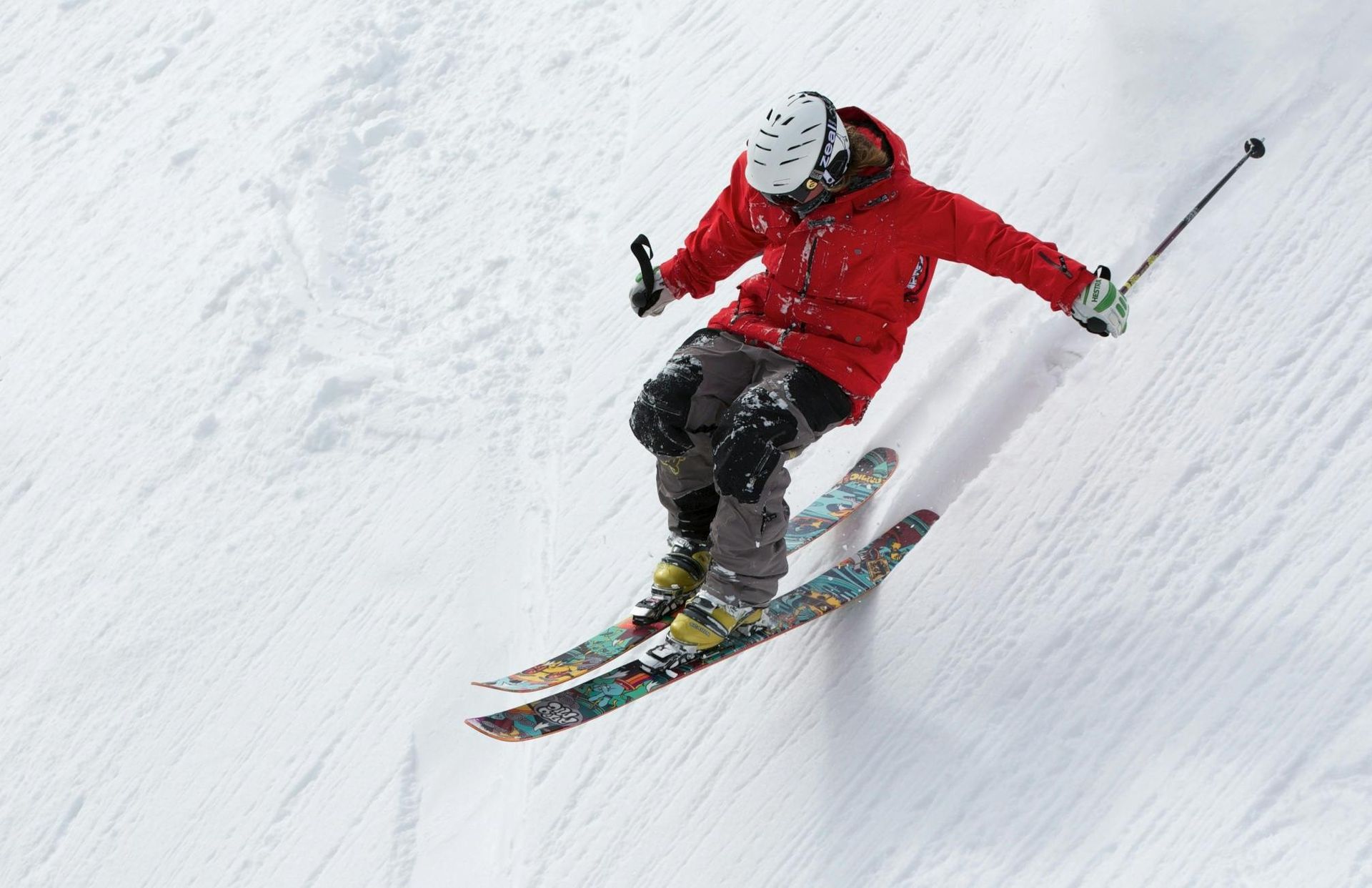 Skier in a red jacket carving down a snowy slope, skis parallel, holding poles, white helmet.