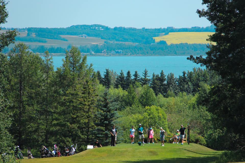 Golfers on a green overlooking a lake and distant landscape, surrounded by trees.