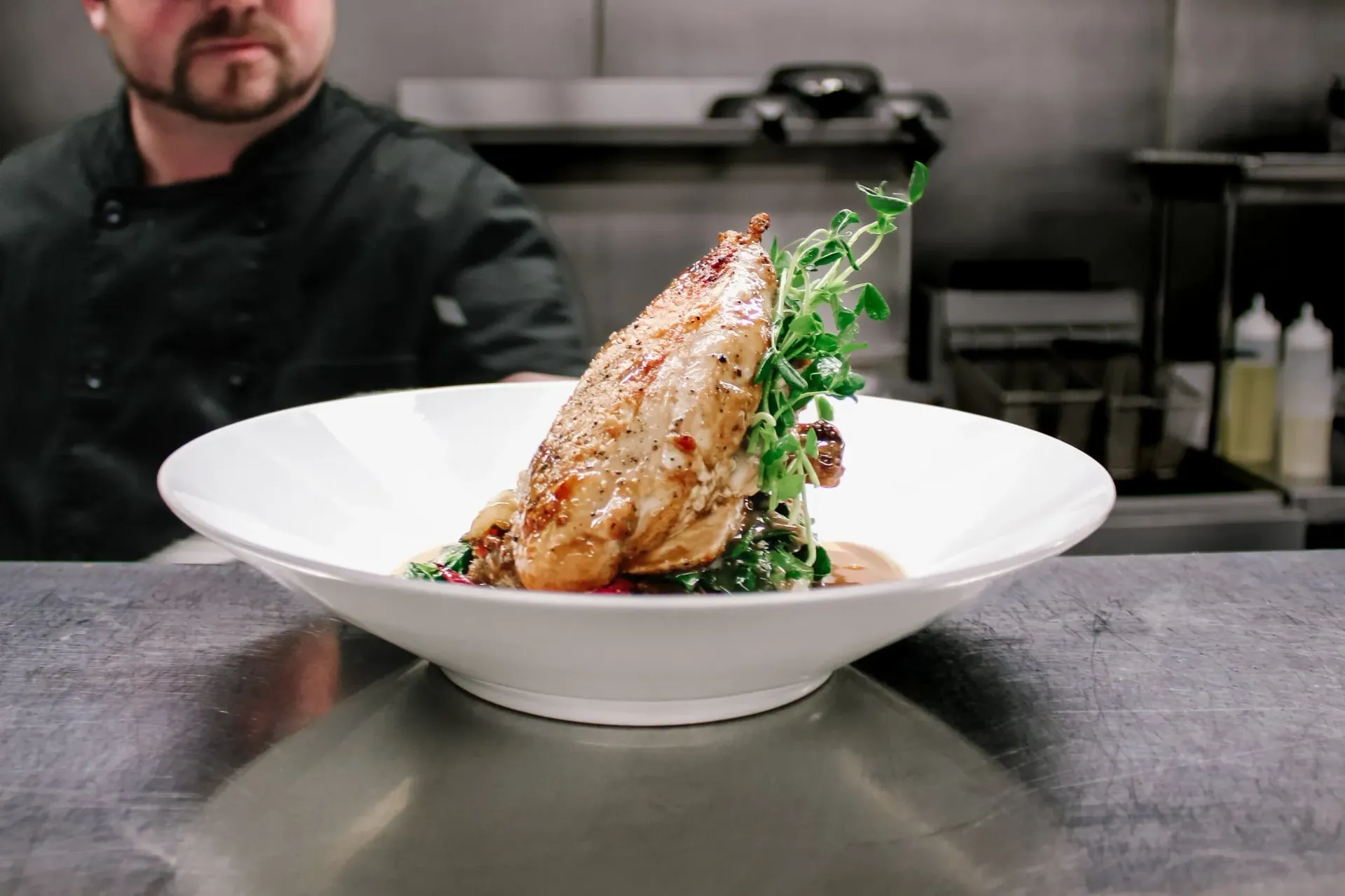 Chef presenting plated chicken dish with greens, on a stainless steel counter.