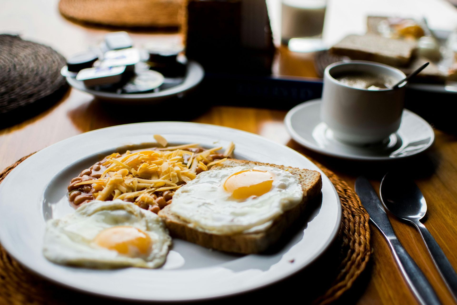 Breakfast plate with fried eggs, beans, cheese, and toast; coffee cup, butter pats, and bread on a wooden table.