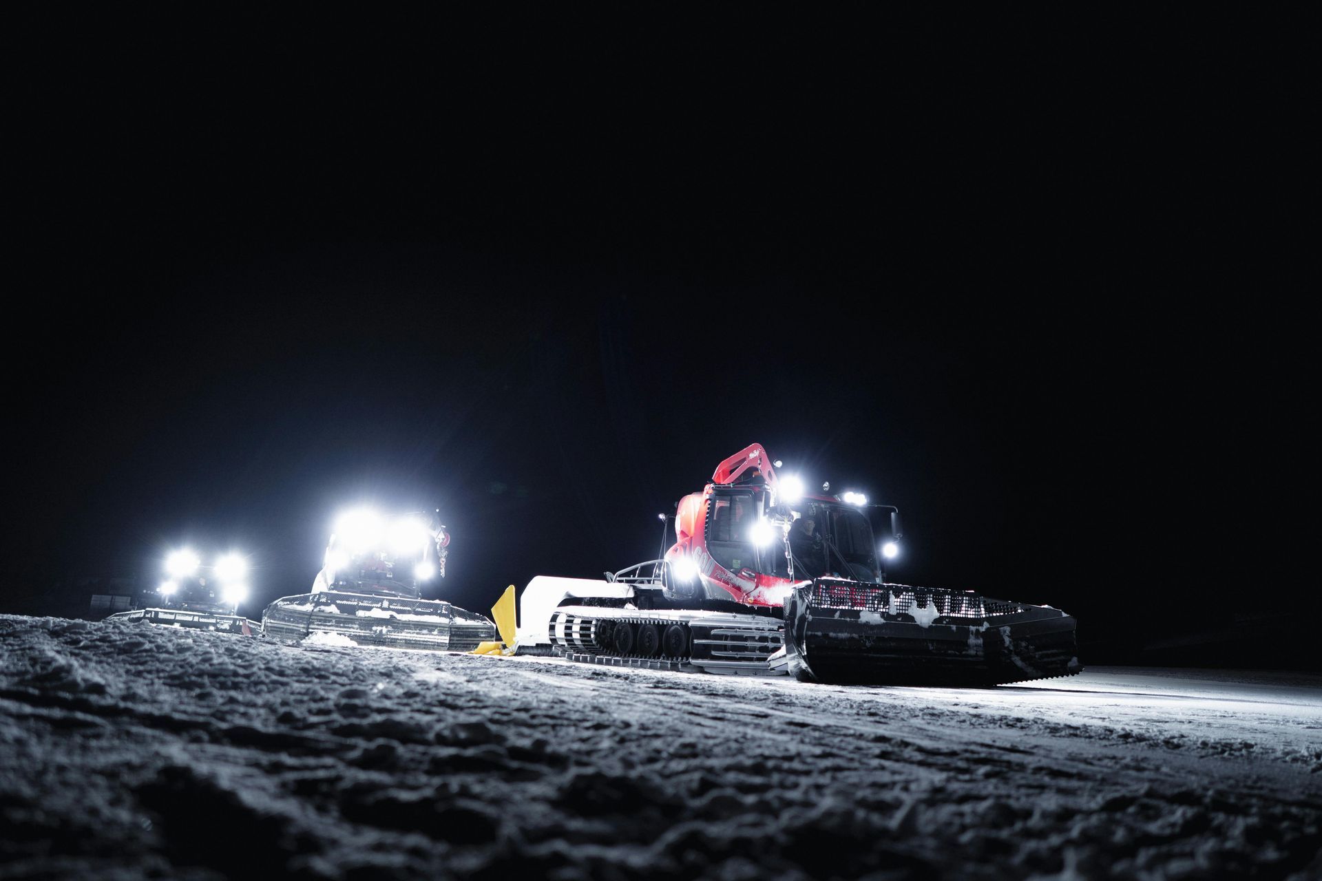 Snow groomers with bright lights working on a snow-covered slope at night.