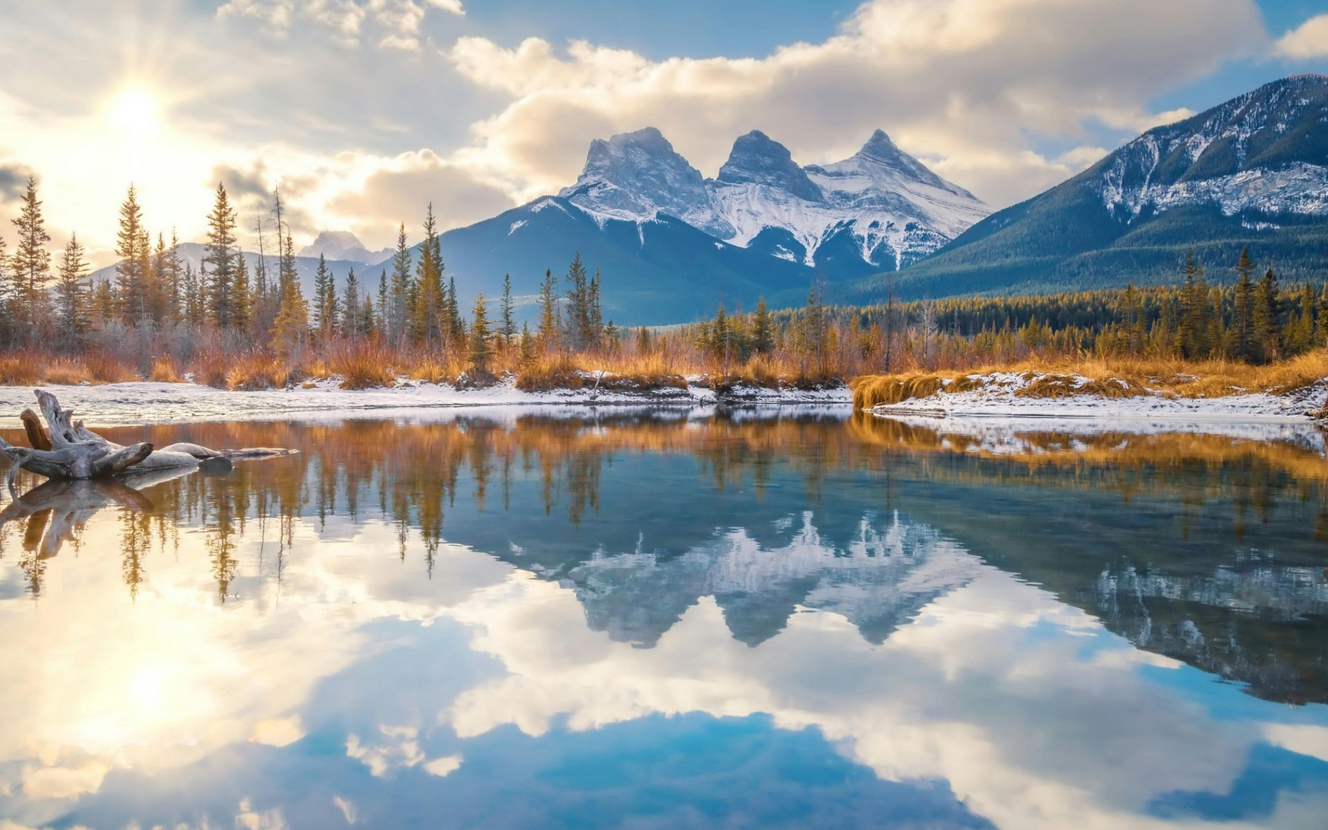 Snow-capped mountains reflected in a calm lake under a bright sky; trees line the shore.