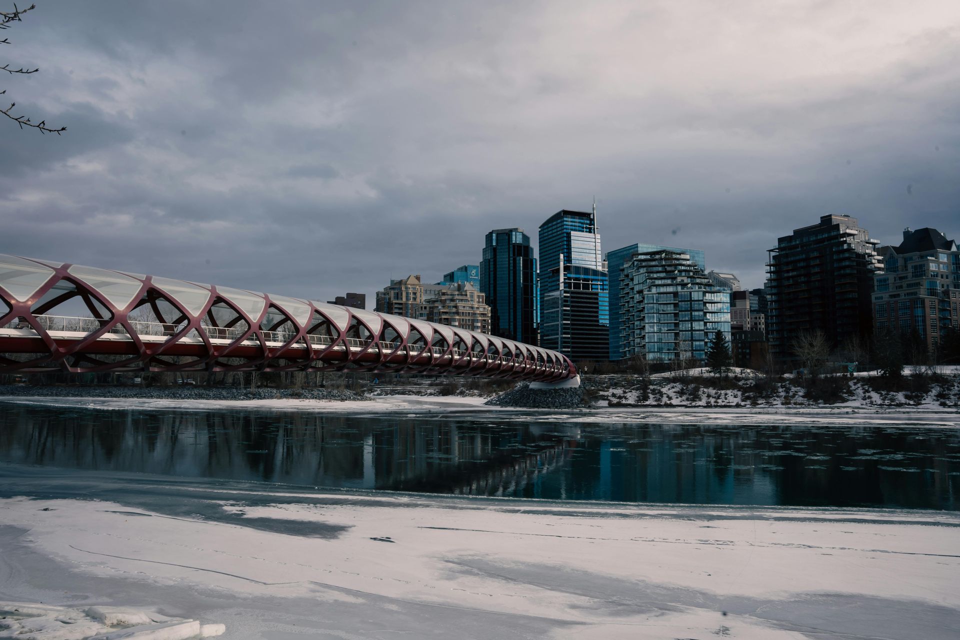A red pedestrian bridge over a partially frozen river, with a city skyline in the background on a cloudy day.