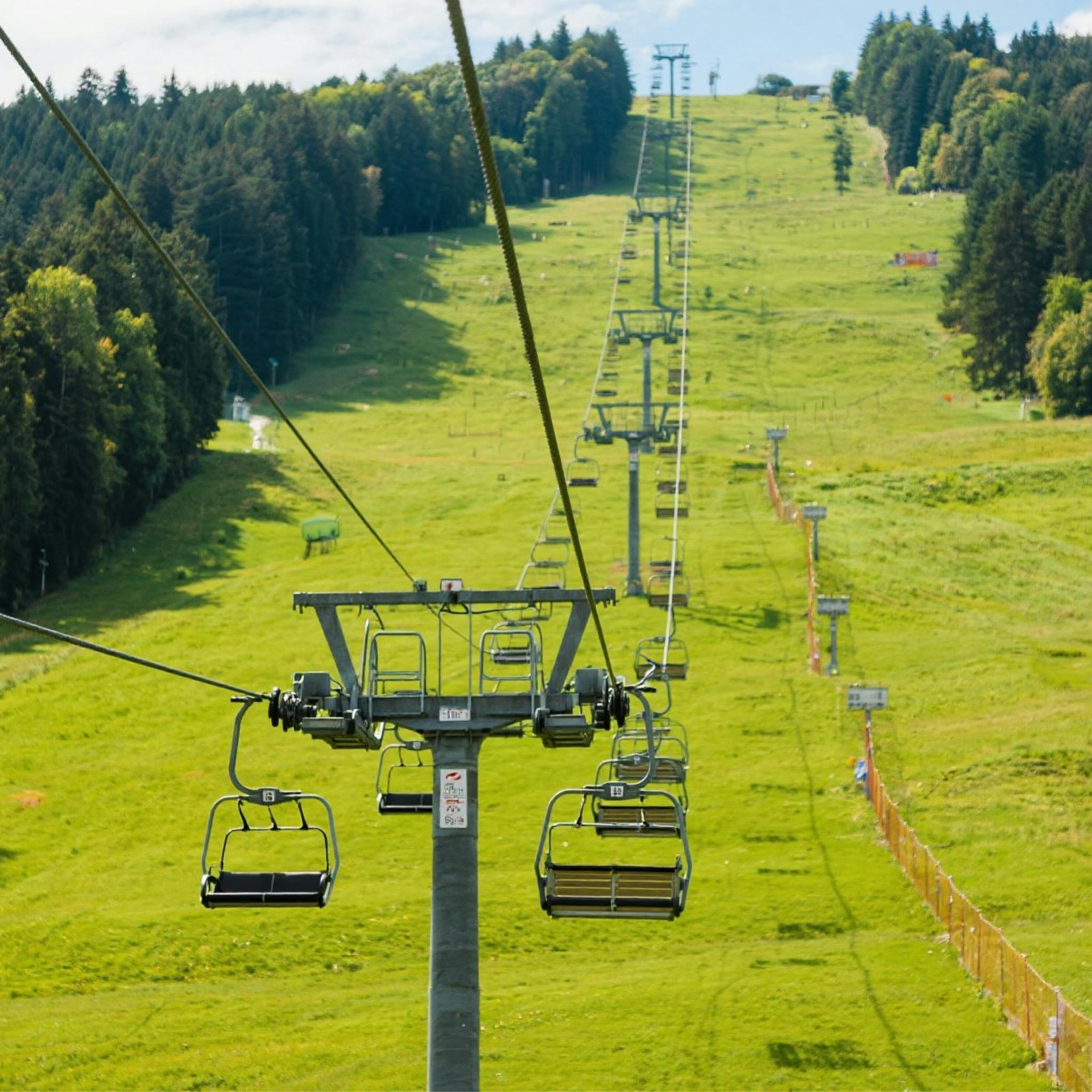 Chairlift ascending a green grassy slope, passing over a ski run, with trees on both sides.