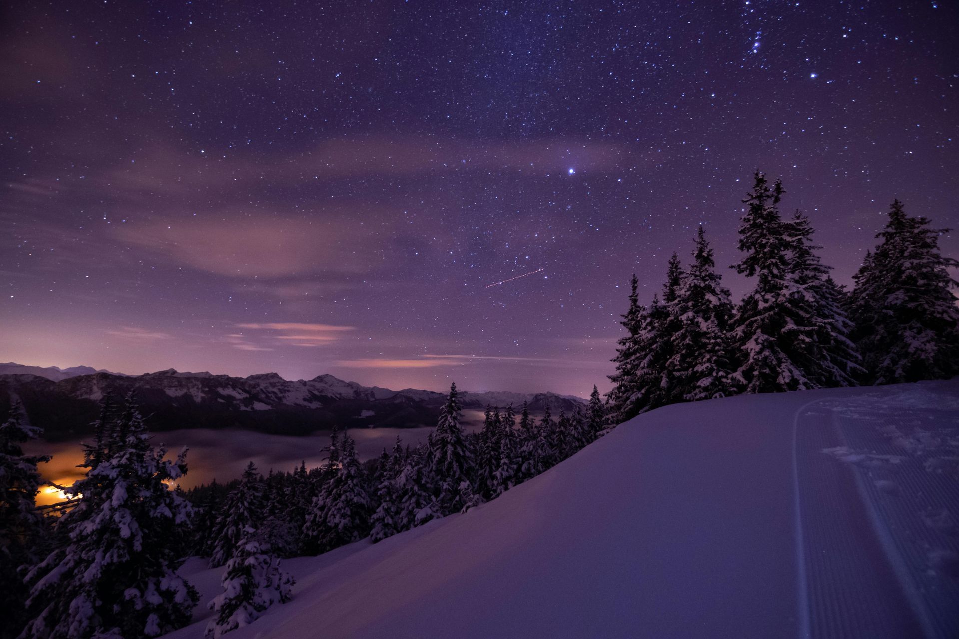 Snow-covered landscape under a starry night sky. Trees silhouetted against purple hues, lake below.