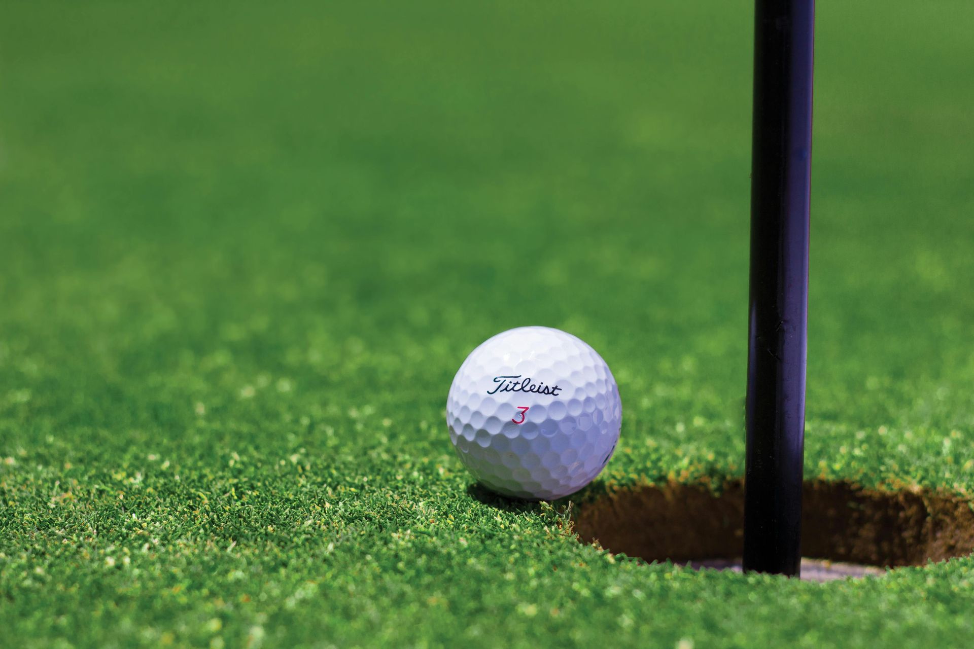 Golf ball near hole with black flag on green grass.