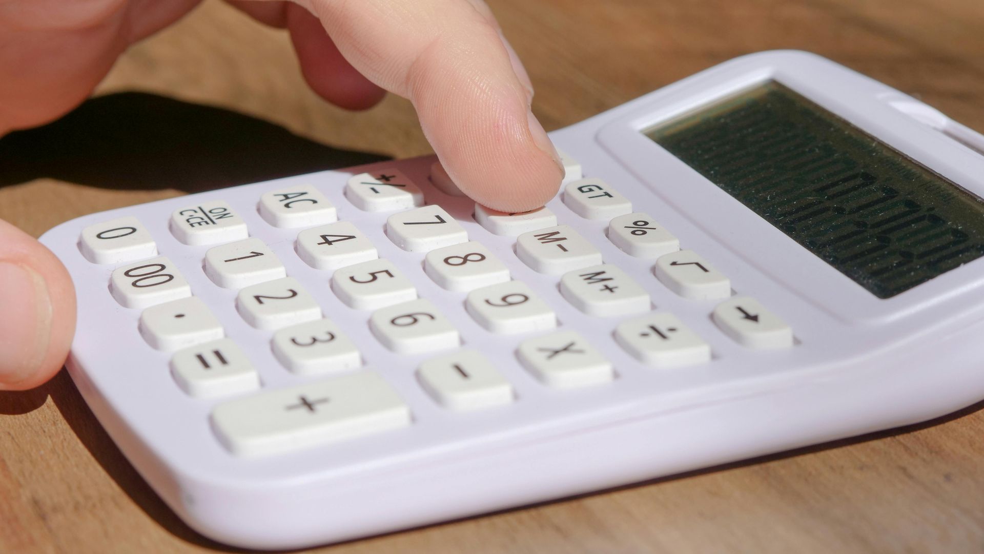 Finger pressing button on a white calculator with a solar panel on a wooden table