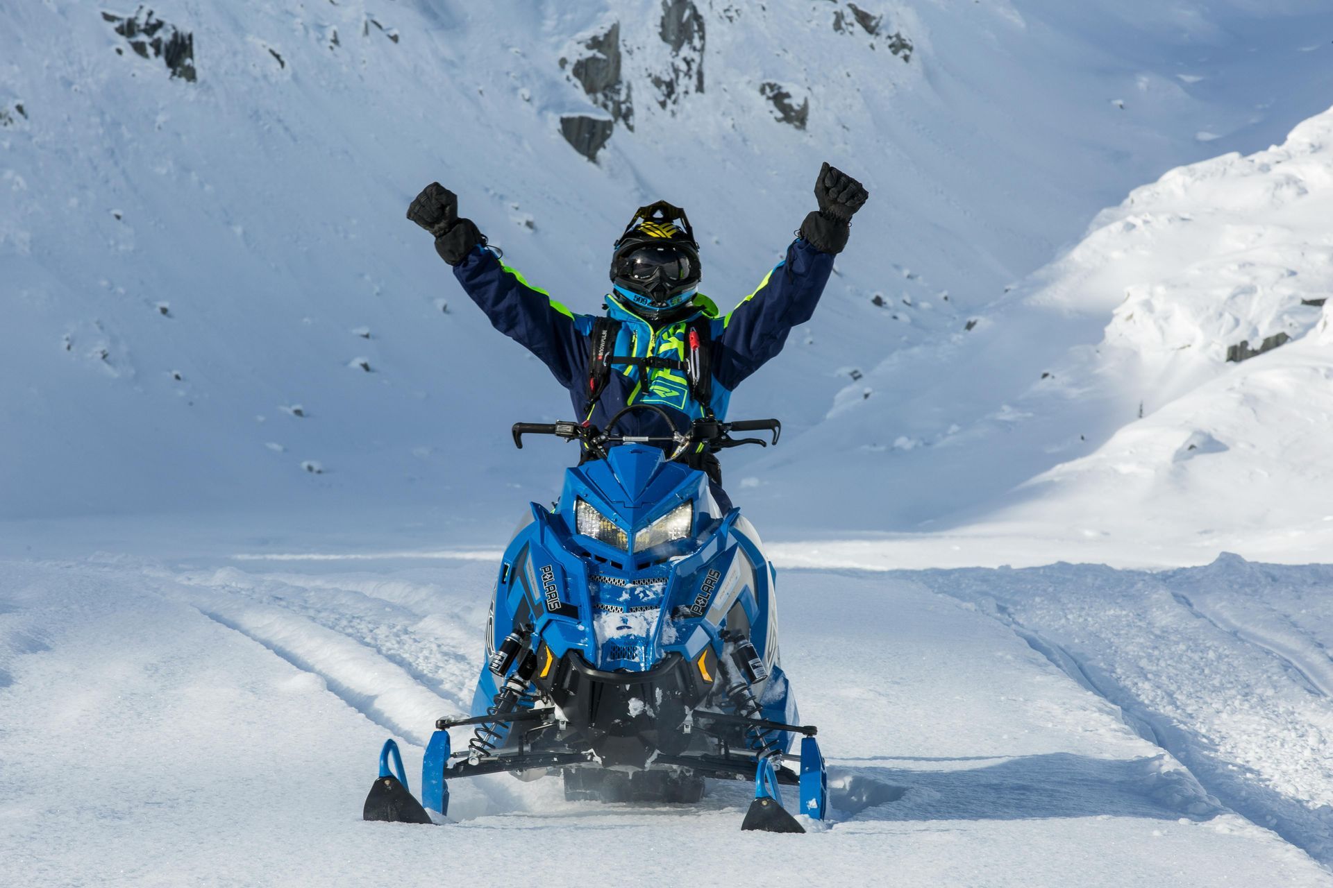 Snowmobiler in blue snowmobile with arms raised in victory, snowy mountain backdrop.