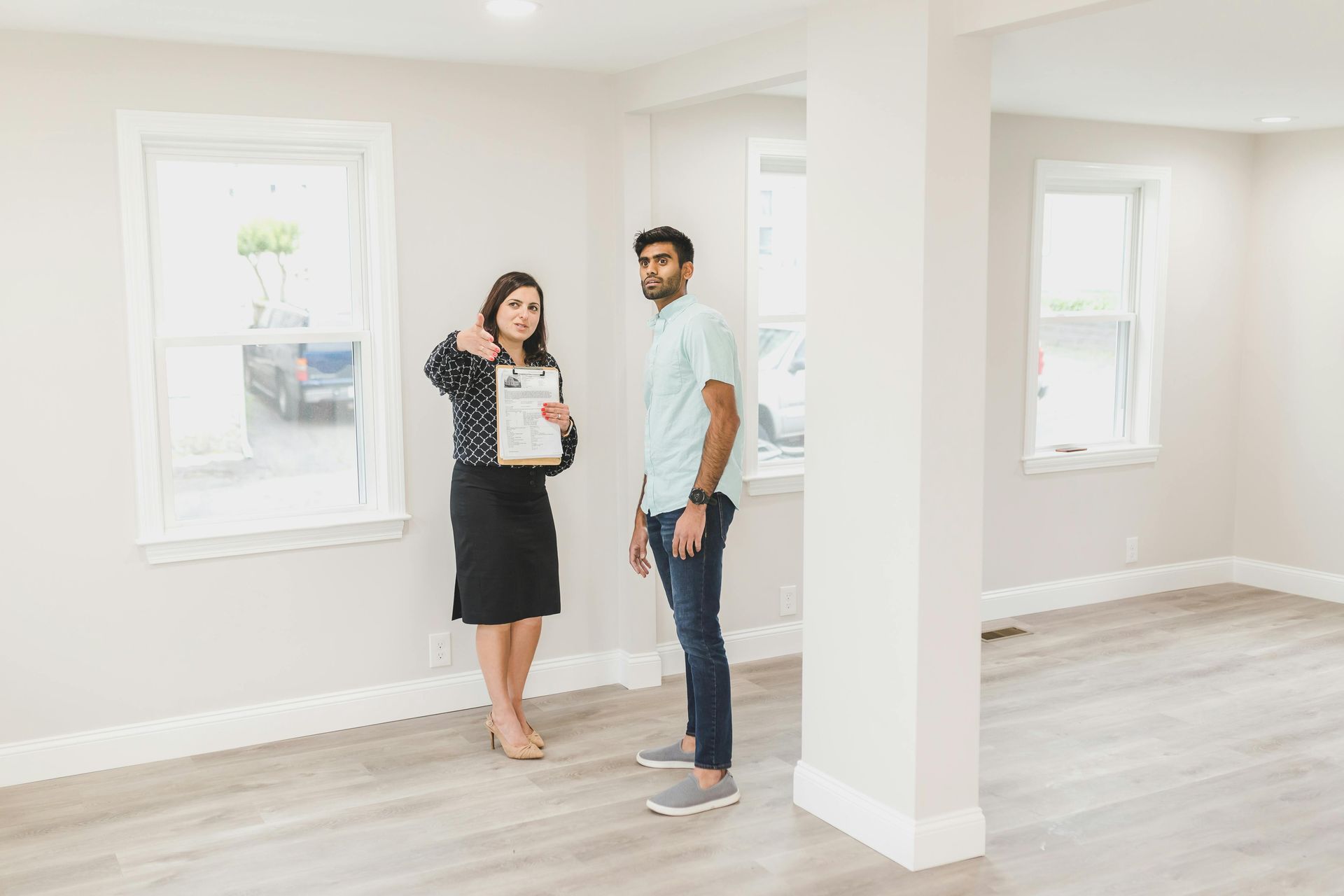 Woman in a black skirt and man in a blue shirt stand in an empty room looking at the camera.
