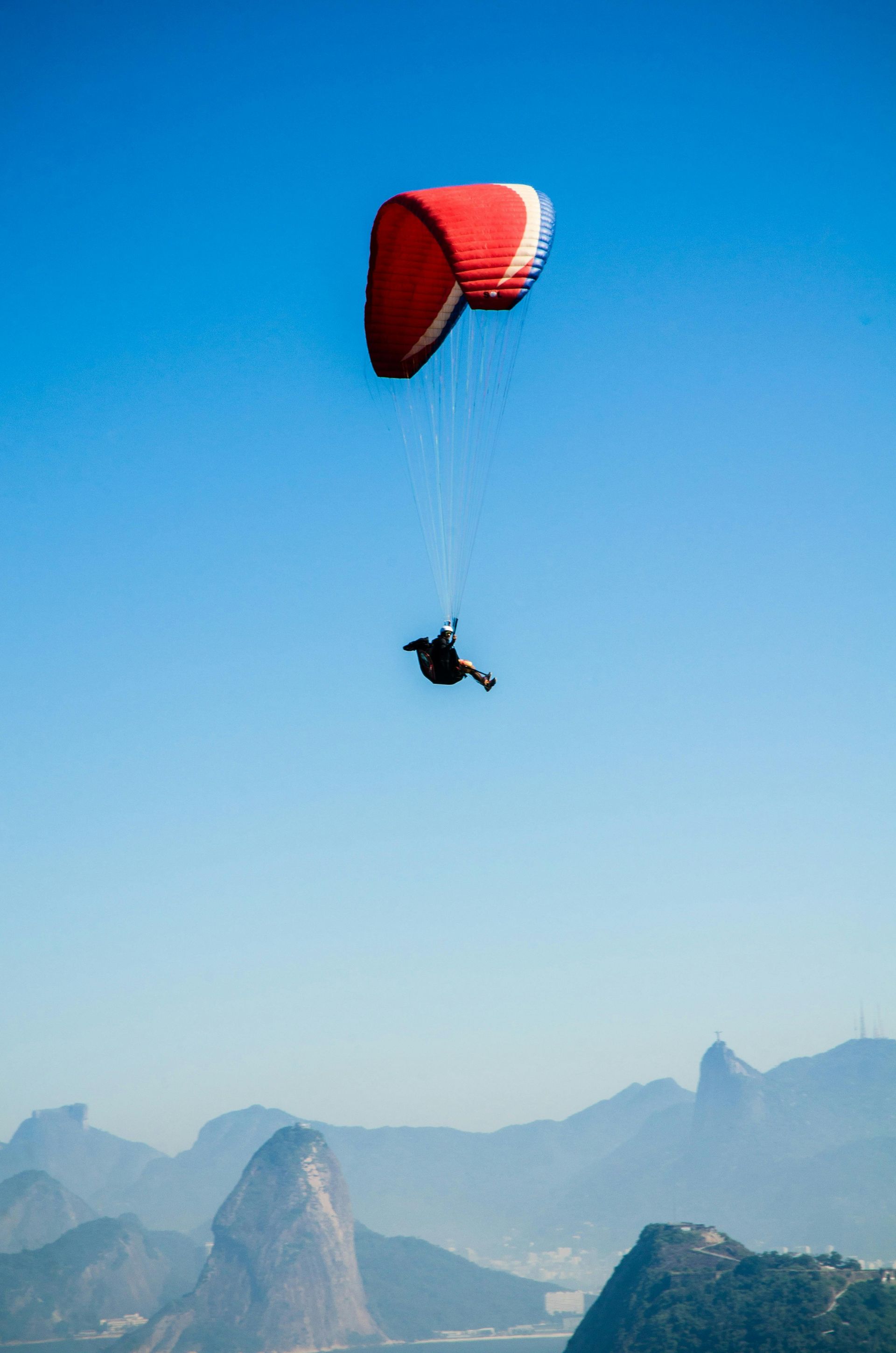Paraglider soaring over mountains under a clear blue sky; red and white wing.