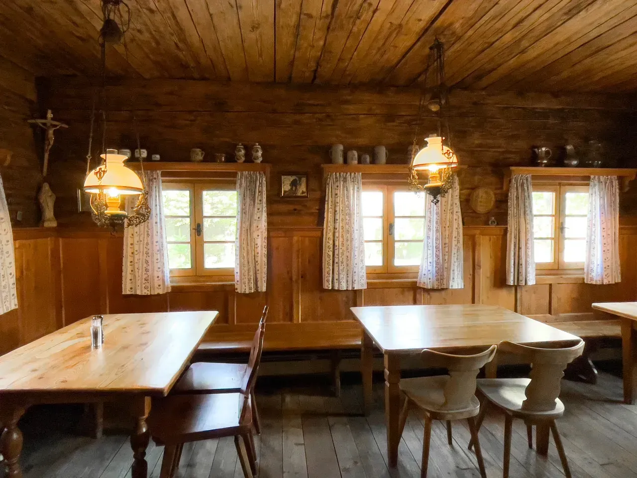Wooden interior of a rustic dining room with tables, chairs, windows with curtains, and hanging lamps.