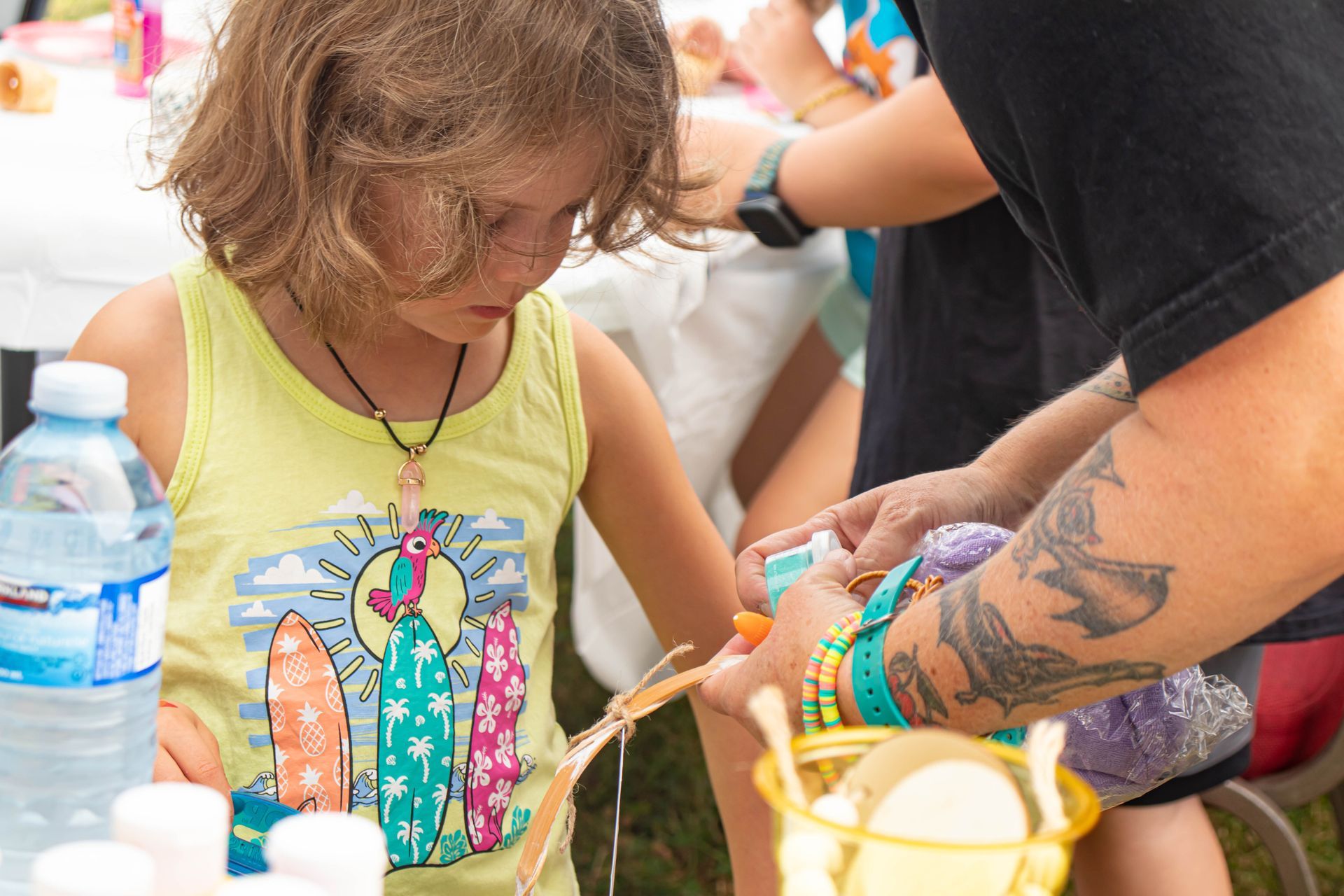 Child in a green shirt painting a colorful craft while an adult assists nearby at a table.