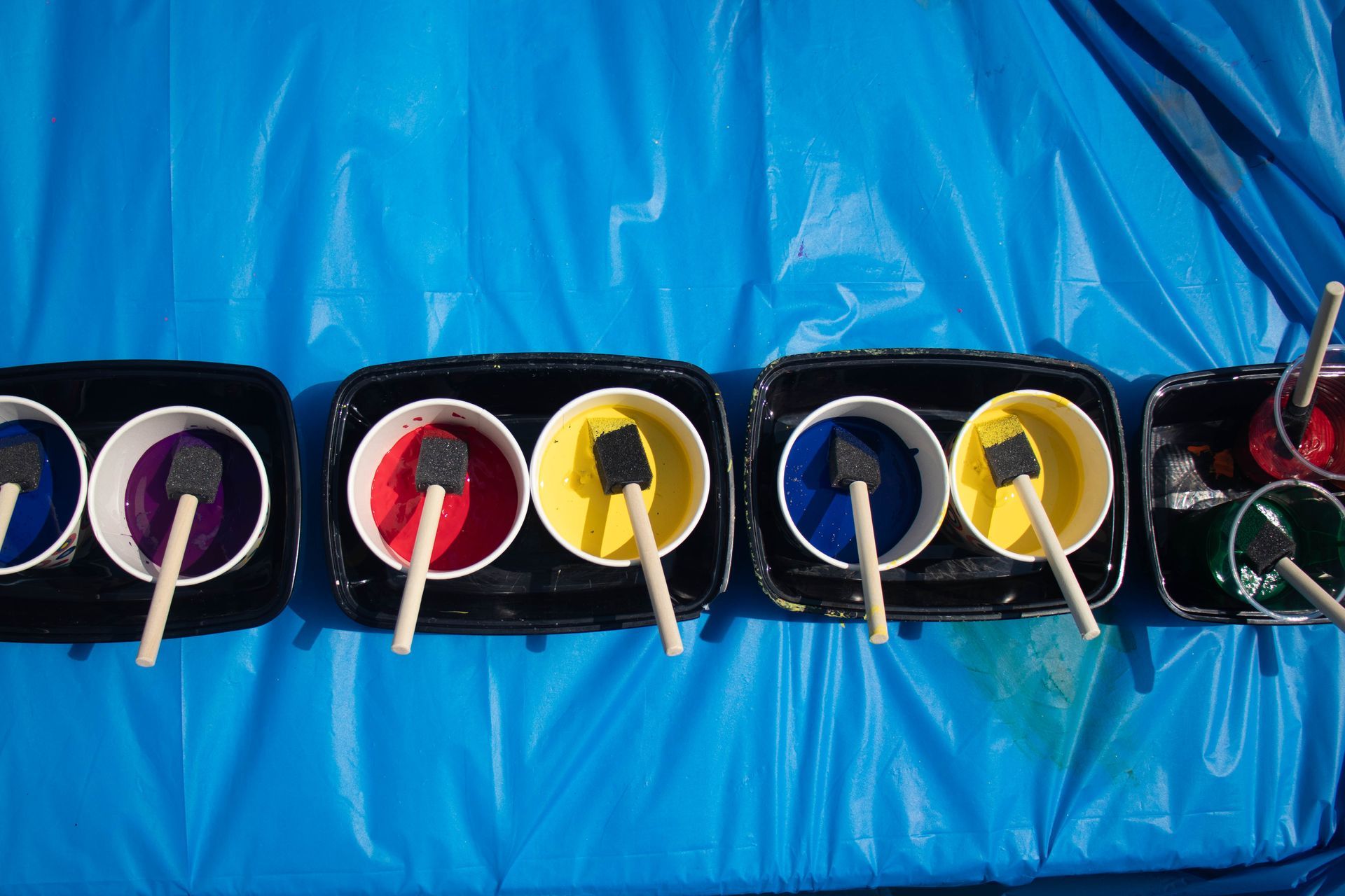 Colorful paint cups with brushes laid out on a blue plastic sheet, likely for a craft or painting activity.