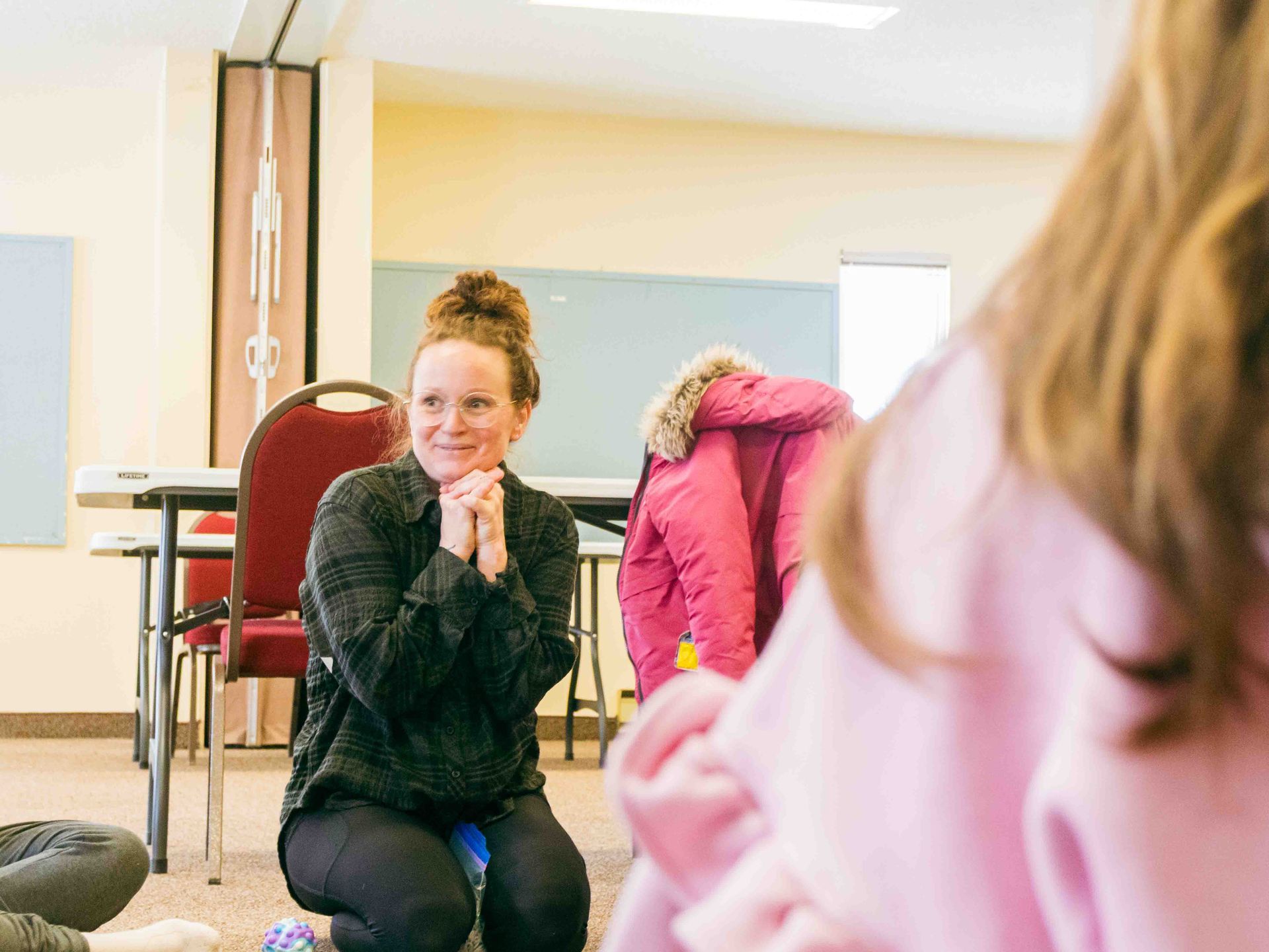 Group activity in a bright room, with a woman sitting on the floor and others nearby in coats and pink clothing