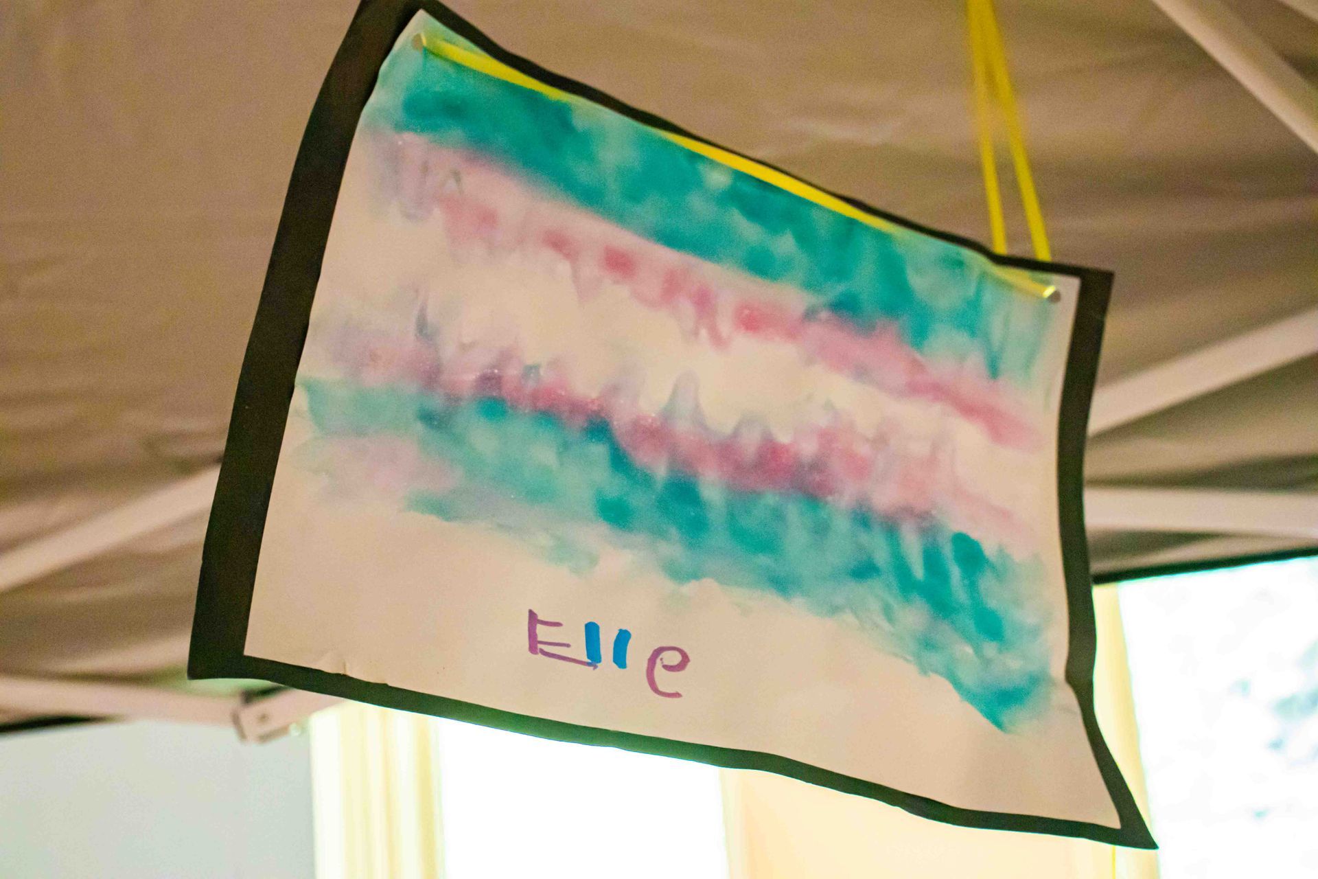 Colorful handmade flag with pink, teal, and white stripes hanging indoors.
