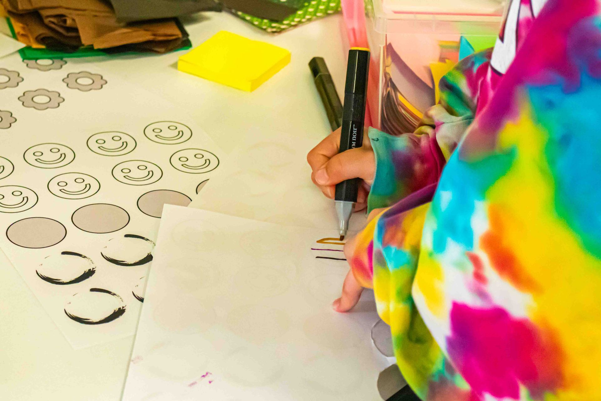 Person in a tie-dye hoodie drawing smiley faces on paper at a desk with markers and notes.