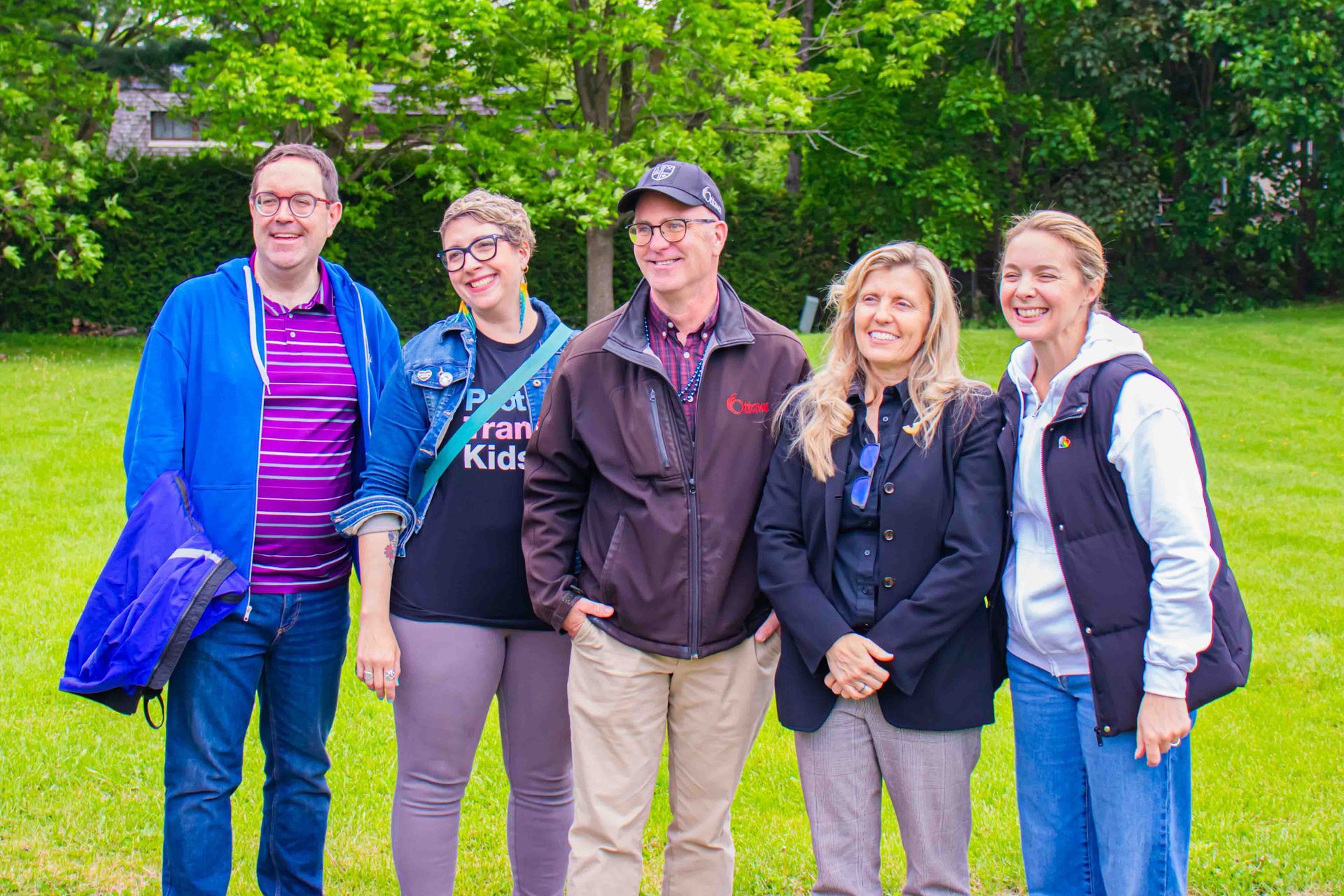Five people standing together outdoors on grass with trees in the background, smiling at the camera.