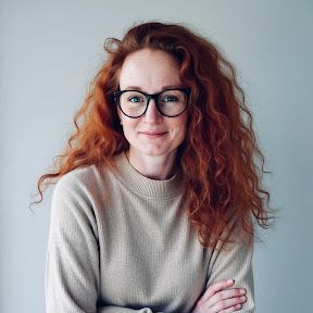 Portrait of a woman with curly red hair and glasses, wearing a beige sweater, against a light background