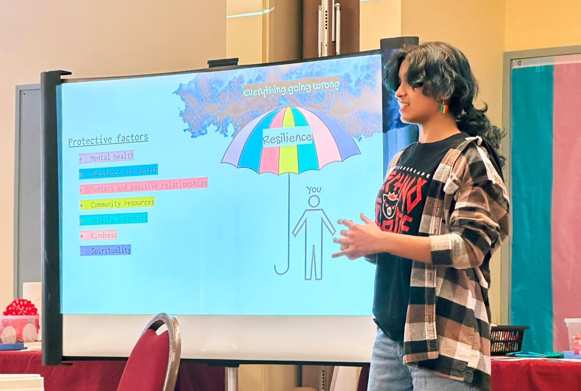 Person presenting beside a colorful slide on a large screen in a classroom or meeting room.