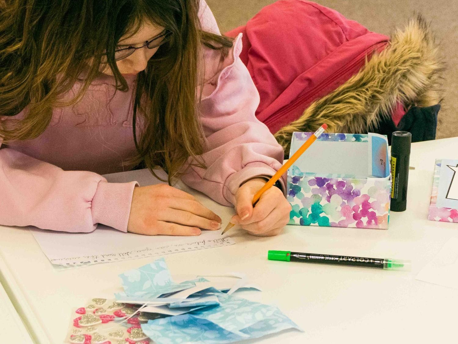 Person drawing at a table with colored pencils and craft supplies, leaning over a colorful collage.