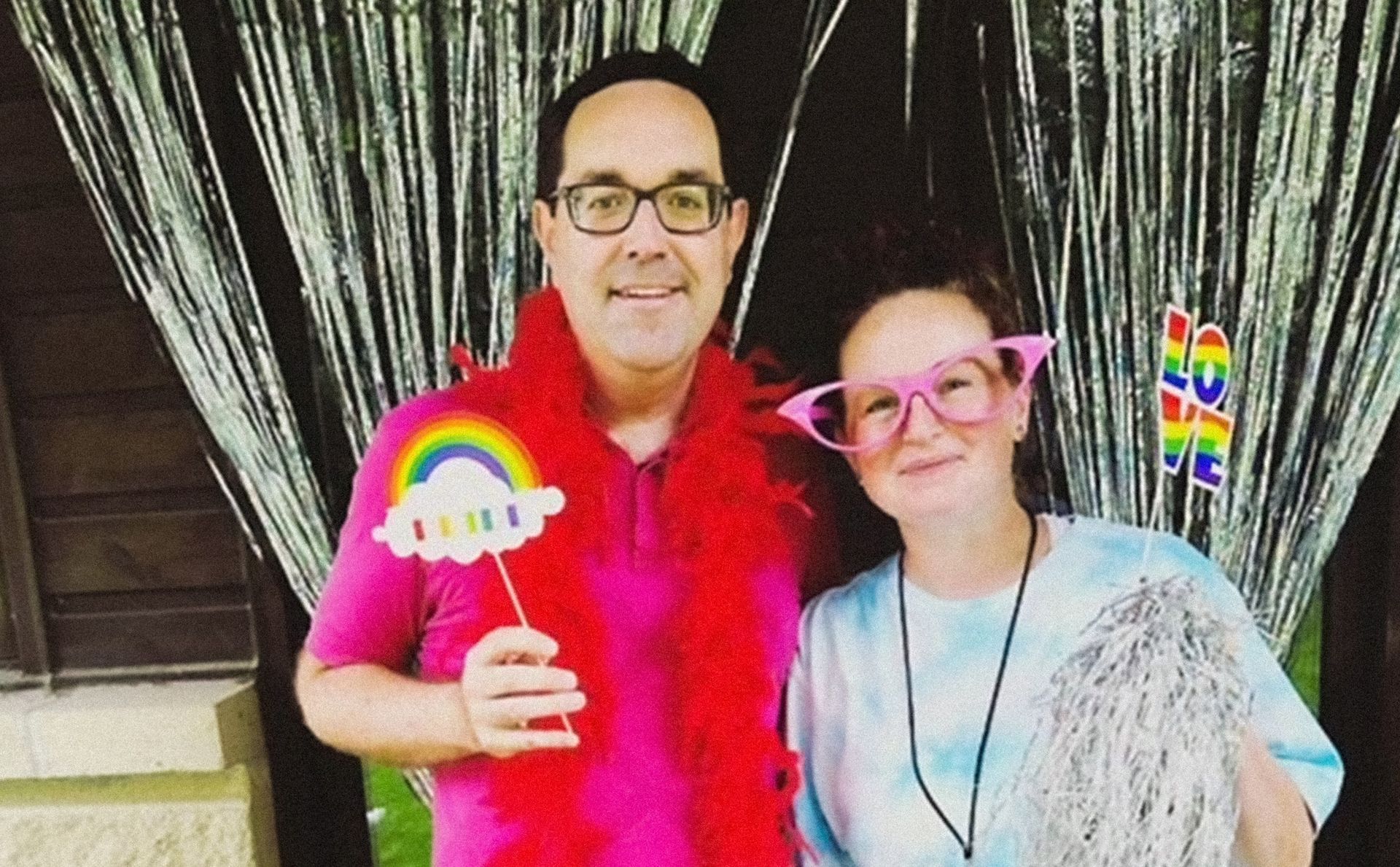 Two people posing indoors, holding rainbow props in front of black curtains, smiling at the camera.