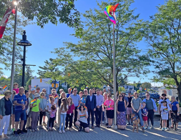 Large group posing outdoors in a sunny park with trees, flags, and a blue sky