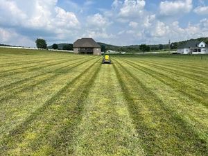 Mowed grassy field with parallel tracks, a small yellow mower, and a house under a cloudy sky.