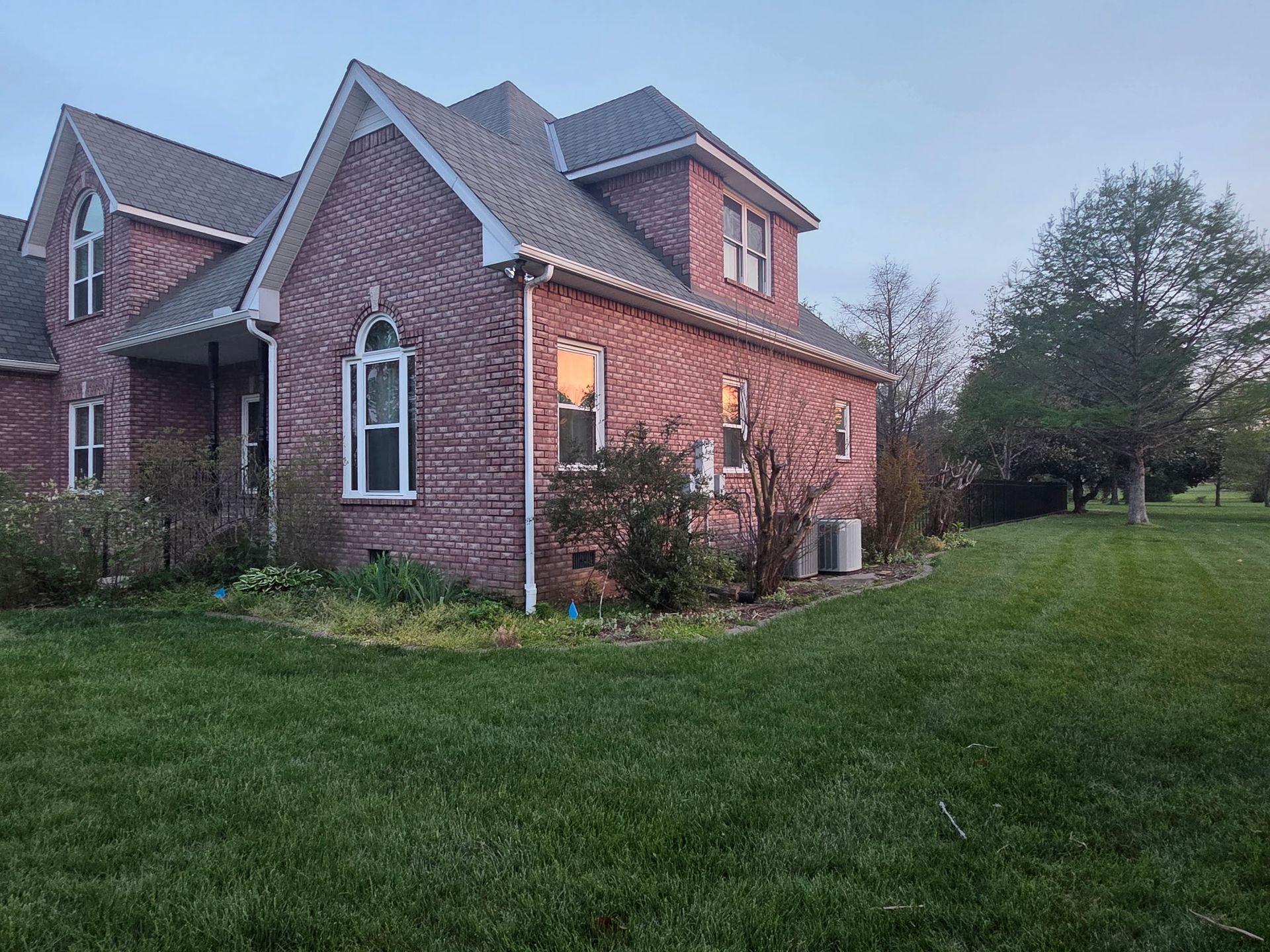 Brick house with lit porch light beside a grassy lawn at dusk
