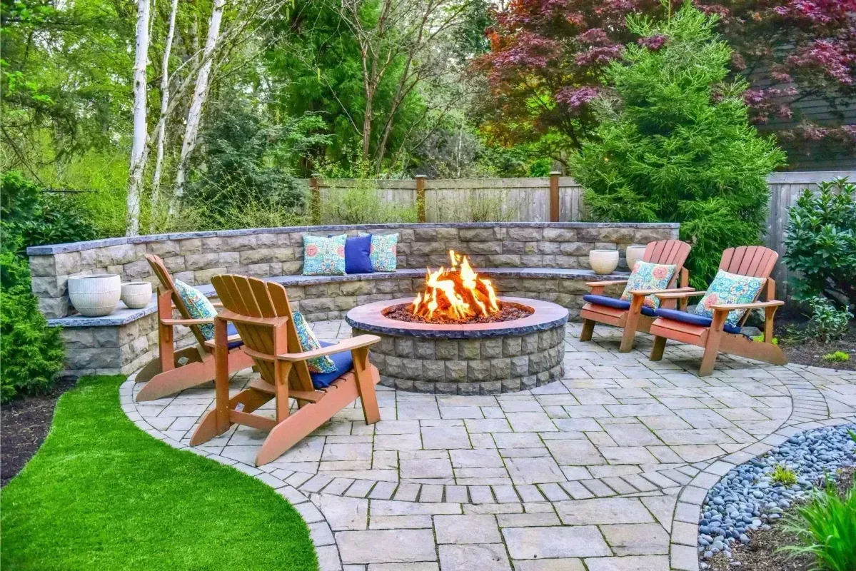 Backyard stone patio with circular fire pit, wooden chairs, and built-in bench seating surrounded by greenery