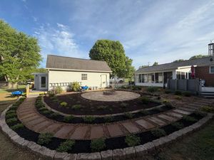 Circular brick garden beds in a landscaped yard with a small cream house and patio seating under a blue sky