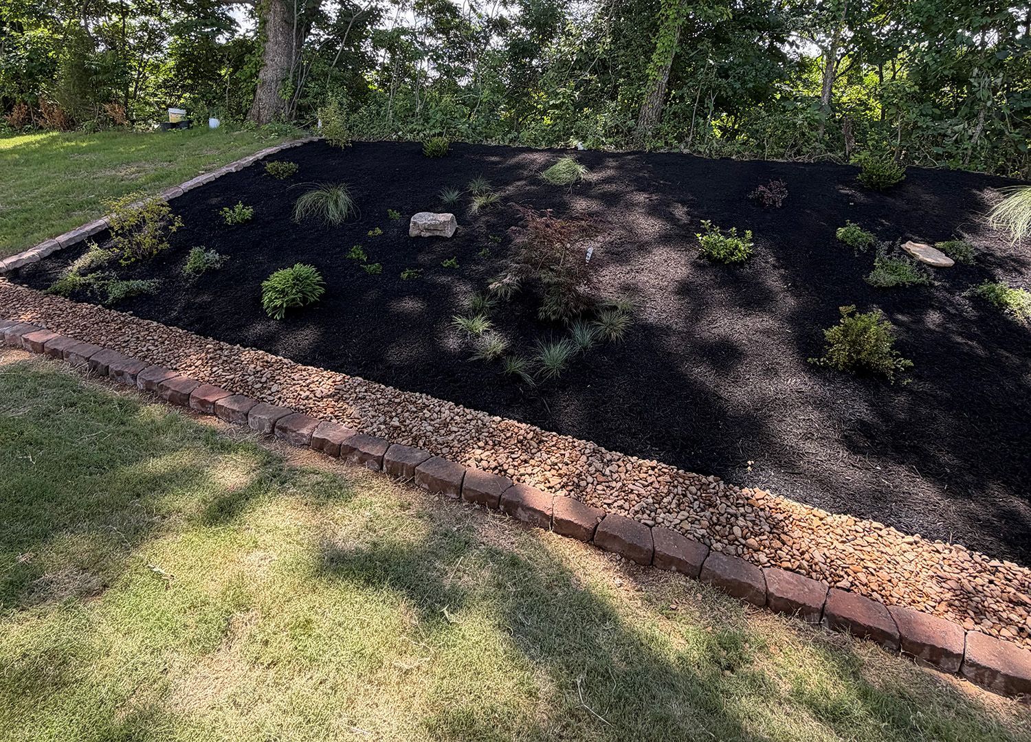 Freshly mulched garden bed bordered by brick, with trees and grass in the background.