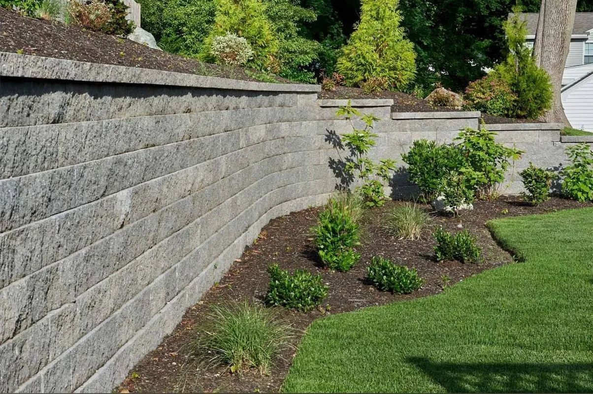 Landscaped garden with stone retaining walls, shrubs, mulch beds, and green lawn on a sloped yard