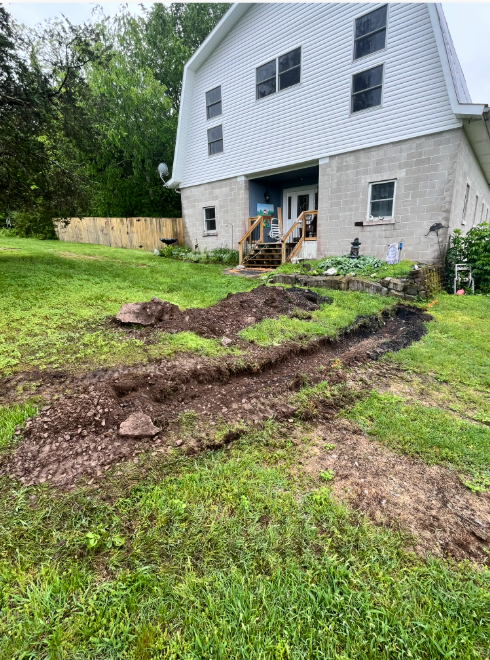 Backyard with fresh dirt trench beside a house and small deck, likely for construction or drainage work