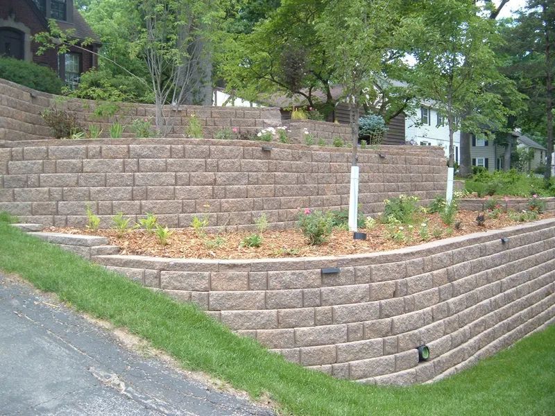 Terraced stone retaining wall with small planted beds beside a sloped driveway and trees
