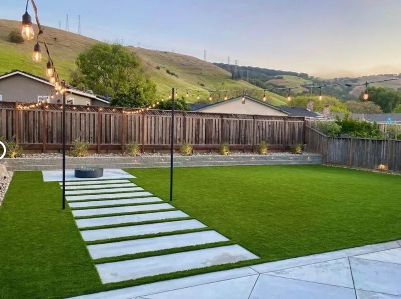 Backyard with green lawn, stepping-stone path, string lights, fence, and hills at sunset