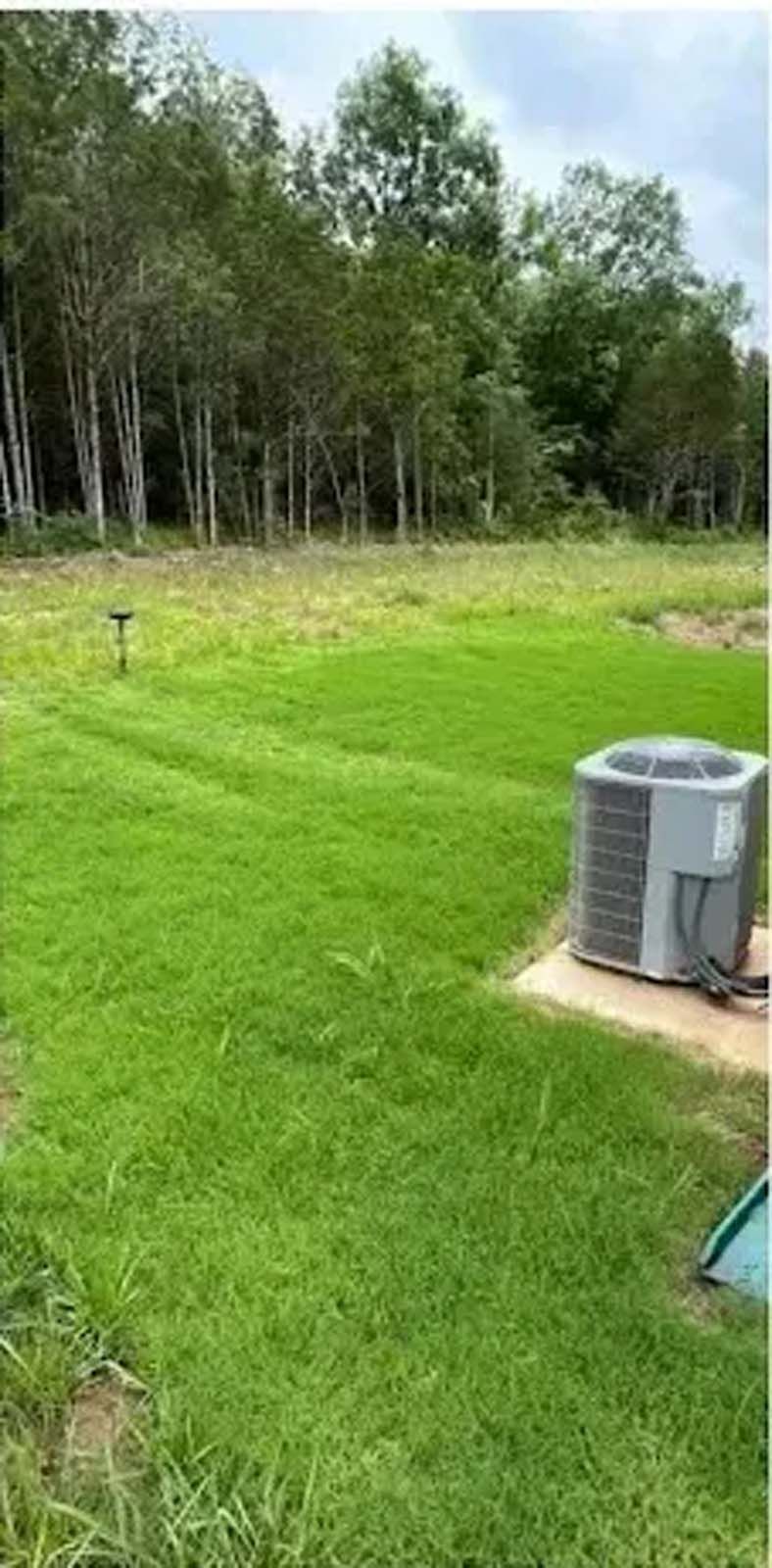Grassy backyard with a small HVAC unit, lawn sprinkler, and trees in the background